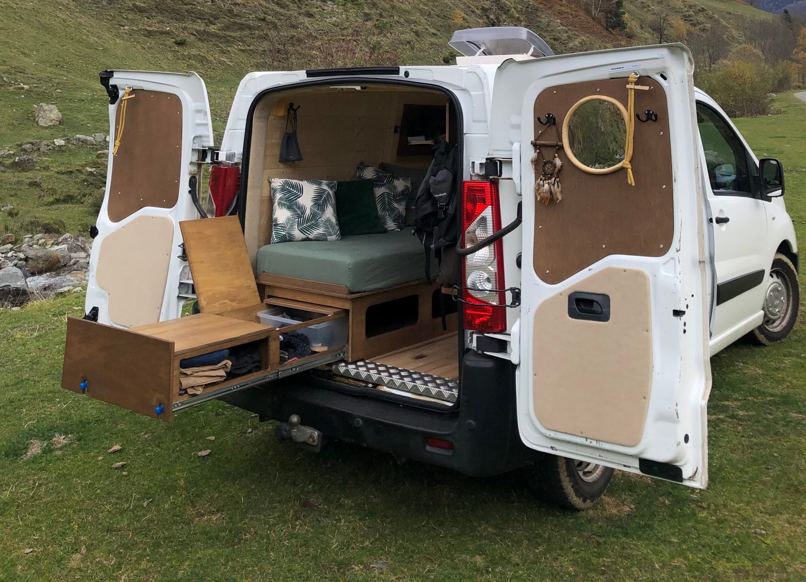 Aménagement intérieur d’un van aménagé avec du bois, disposant d'un lit peigne, d'un coin cuisine, rangements et espace cosy pour la vie en van